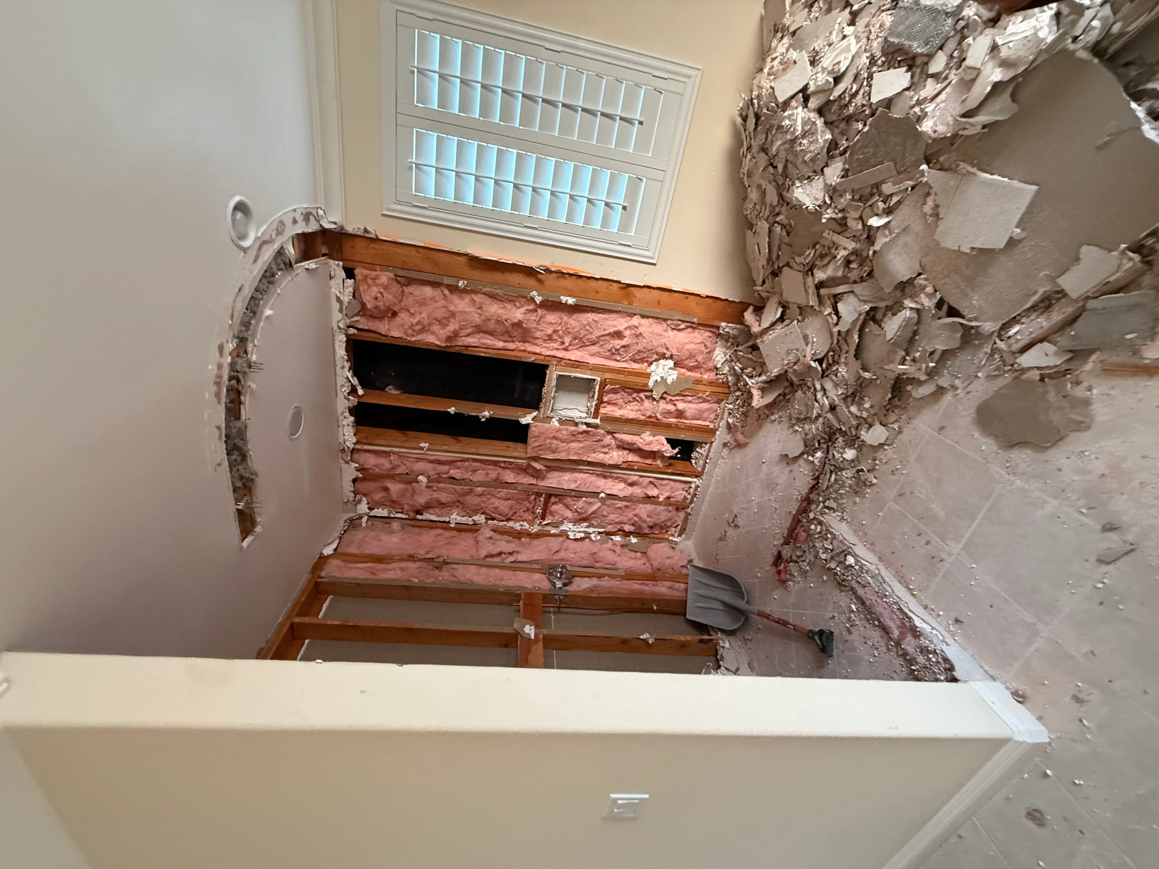 Bathroom wall demolition showing exposed wood framing, pink insulation, and plumbing rough-ins